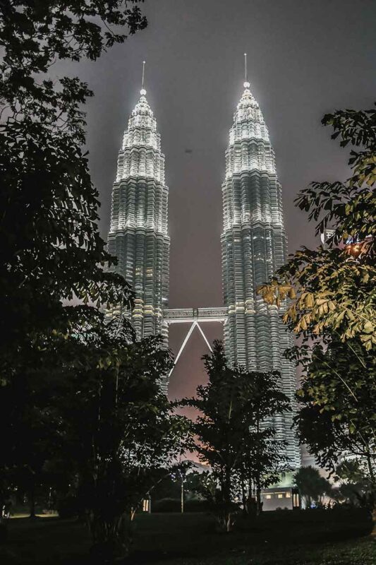 Oświetlone wieże Petronas w Kuala Lumpur nocą, widziane spomiędzy drzew