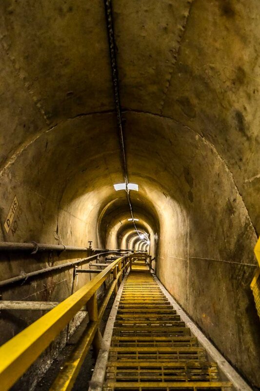 Długi tunel techniczny wewnątrz Zapory Hoovera z metalową podłogą, żółtą balustradą i betonowymi ścianami łukowymi