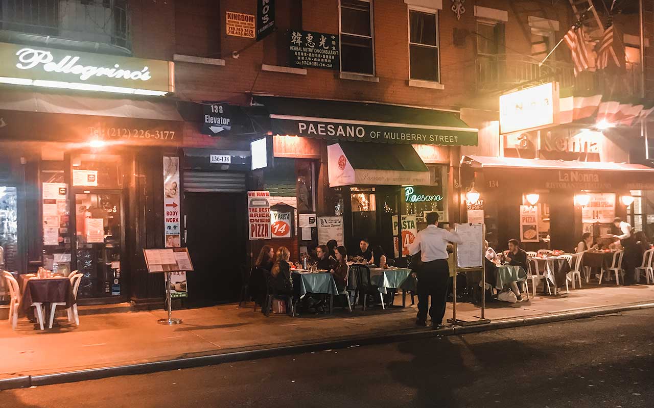 Stoliki na zewnątrz restauracji Paesano of Mulberry Street i La Nonna w Little Italy oświetlone ciepłym światłem wieczoru.