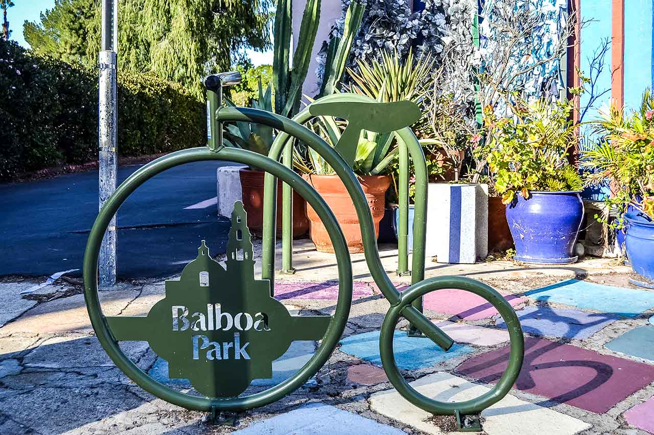 Metalowy stojak na rowery w kształcie stylizowanego bicykla z napisem Balboa Park, ustawiony na tle kolorowego chodnika i roślin w ogrodzie